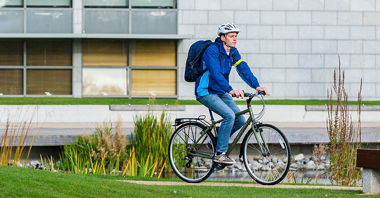A young male student wearing a blue jacket rides a bike beside the Daedalus building on campus.